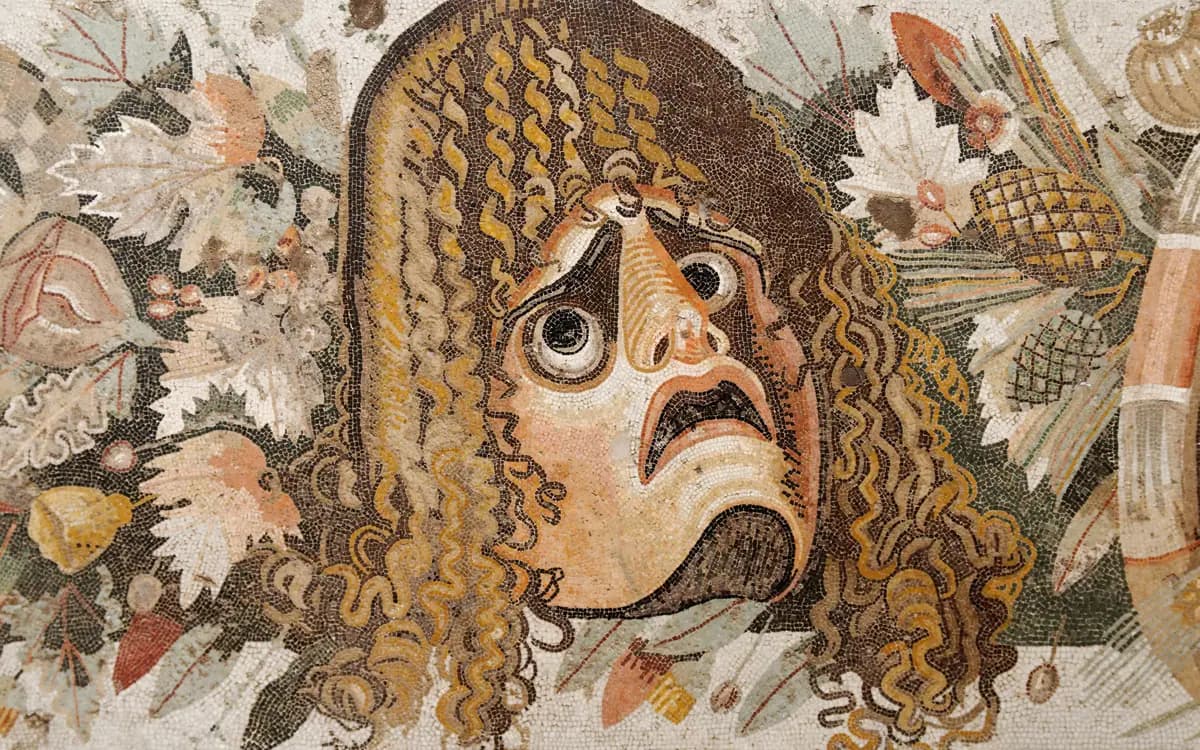 Ancient Roman floor mosaic of a tragic theatre mask with wide eyes, an open mouth and long curling hair, surrounded by leaves and fruit.