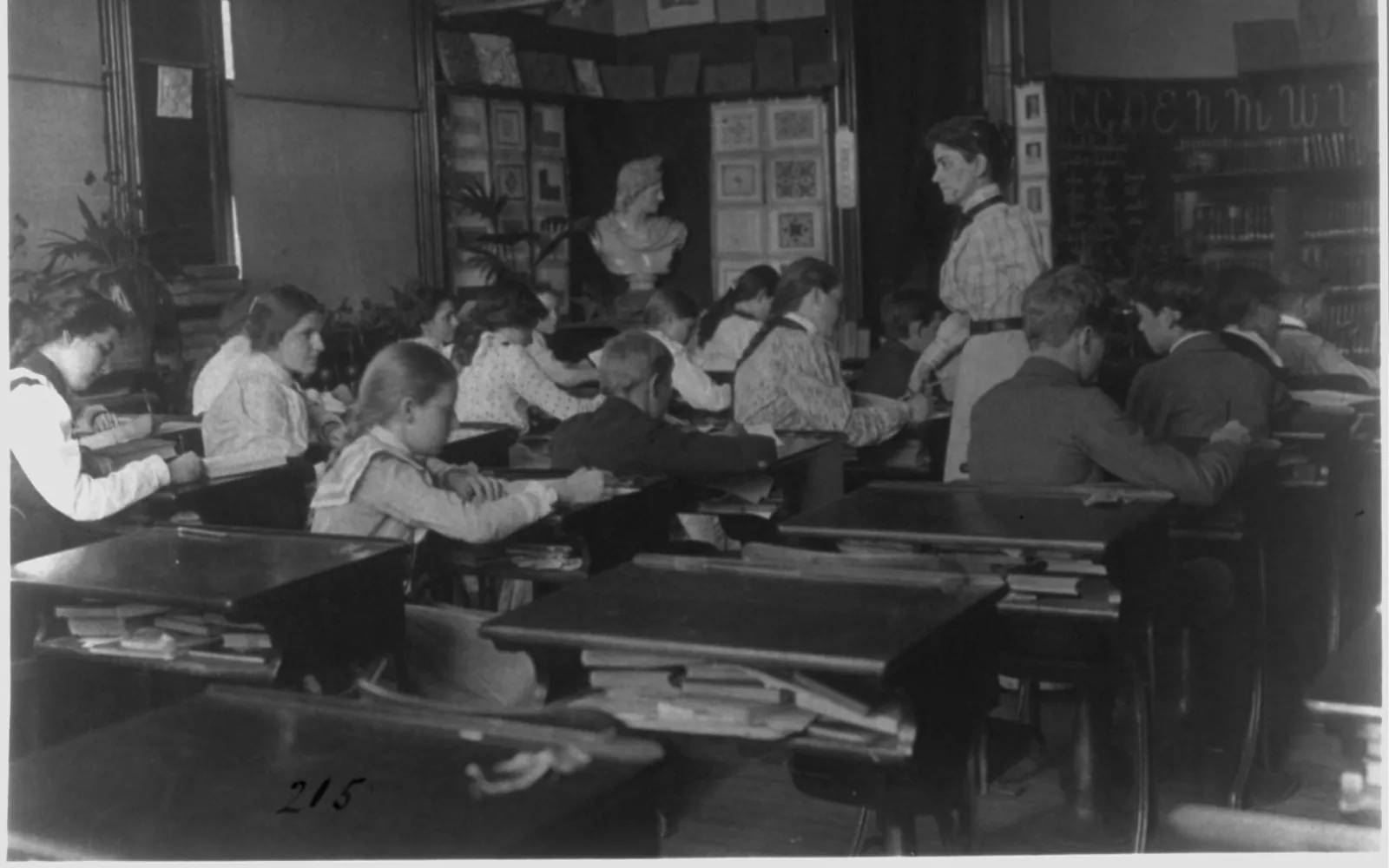 Black and white photograph of a school classroom around 1899, with boys and girls writing at wooden desks while a teacher in a striped blouse stands among them, and a plaster bust and pictures on the wall behind.