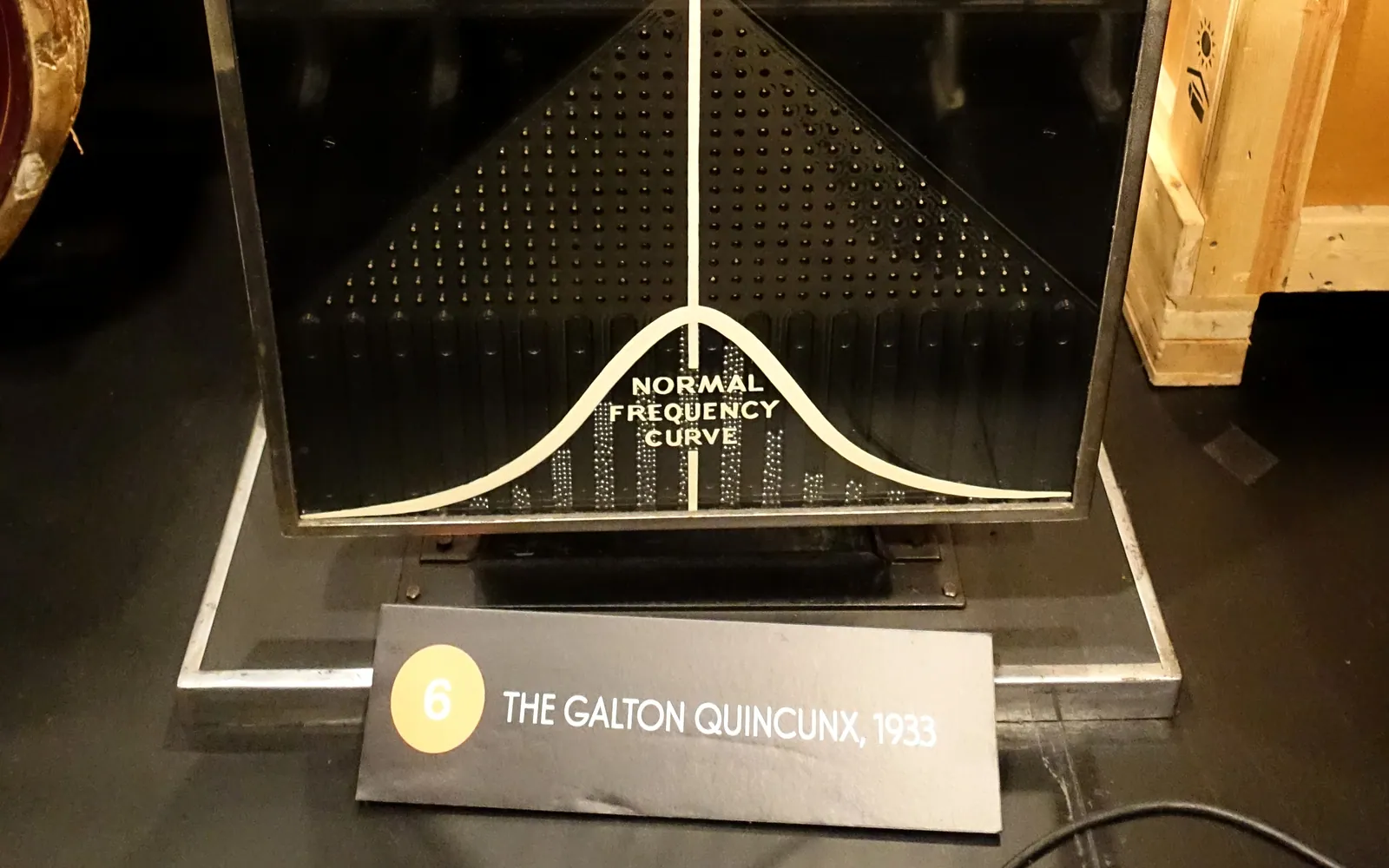 A 1933 Galton quincunx in a museum: a black board of pins above vertical slots where small balls pile up under a painted bell curve labeled Normal Frequency Curve