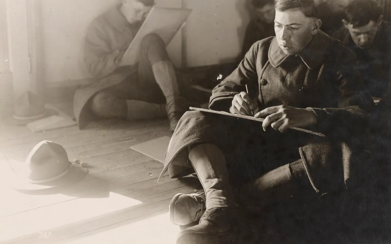 Sepia photograph of young American soldiers in greatcoats sitting on a wooden floor, writing answers on boards resting on their knees during a group intelligence test